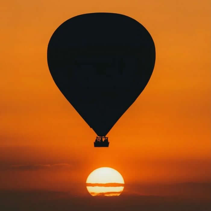 Hot air balloon silhouette flying across a bright orange sunset sky with the sun near the horizon creating a dramatic minimalist sunset scene.
