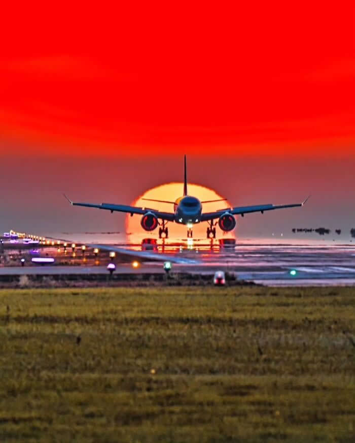 Airplane landing on runway aligned with the setting sun against a dramatic red sunset sky creating a striking aviation silhouette scene.