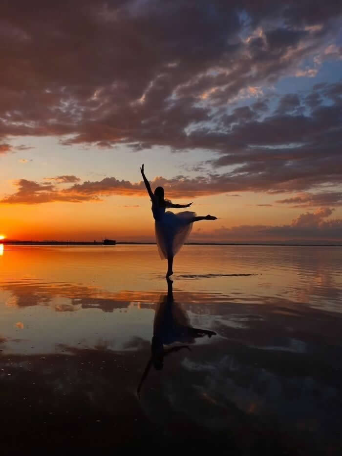 Silhouette of a ballerina dancing on reflective beach water during a golden sunset with dramatic clouds and glowing sky mirrored on the surface.