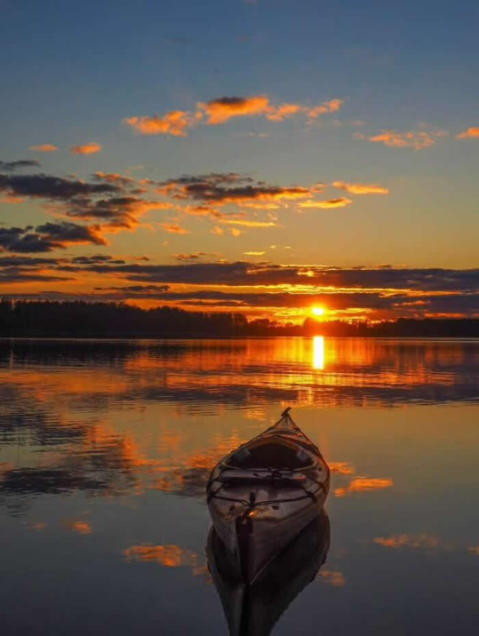 Kayak floating on calm lake reflecting a golden sunset with dramatic clouds and glowing horizon mirrored across the peaceful water surface.