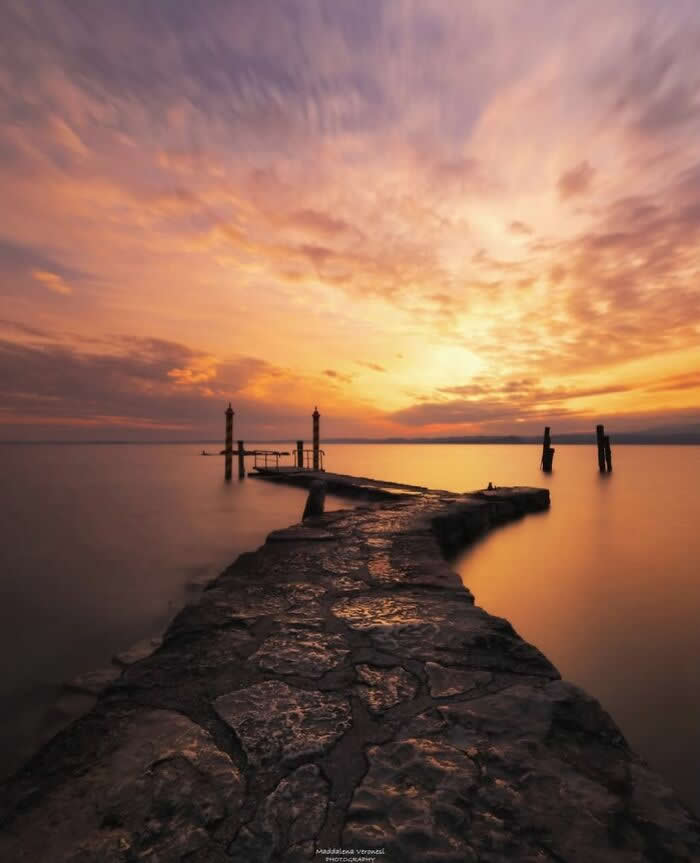 Stone pier extending into calm sea during a glowing golden sunset with dramatic clouds and smooth water reflections at twilight.