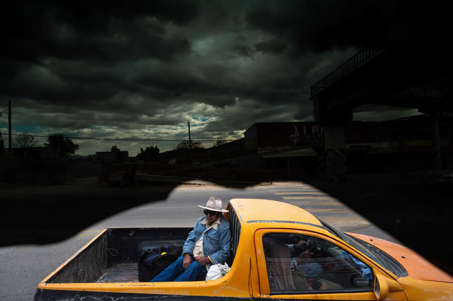Man sitting in the back of a yellow pickup truck framed by dark shapes from torn window film, creating a dramatic illusion resembling storm clouds in a creative street photography composition. - Mexico Street Photography by Tavepong Pratoomwong