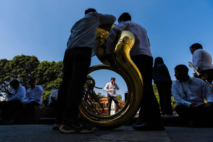 Street musicians gathered outdoors with a large brass tuba in the foreground, framing a man standing in the center, captured with strong contrast between shadows and bright blue sky. - Mexico Street Photography by Tavepong Pratoomwong