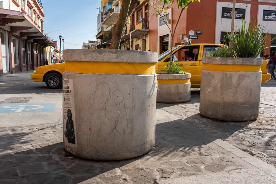 Concrete planters with yellow bands in the foreground align with a yellow taxi passing on a colorful street, creating a layered composition with repeating colors and urban elements in a street photography scene. - Mexico Street Photography by Tavepong Pratoomwong
