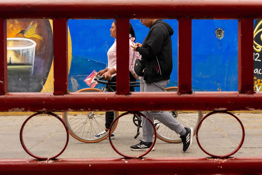 People walking and cycling seen through a red metal frame structure, with circular shapes and a colorful blue wall in the background, forming a layered and geometric street photography composition. - Mexico Street Photography by Tavepong Pratoomwong