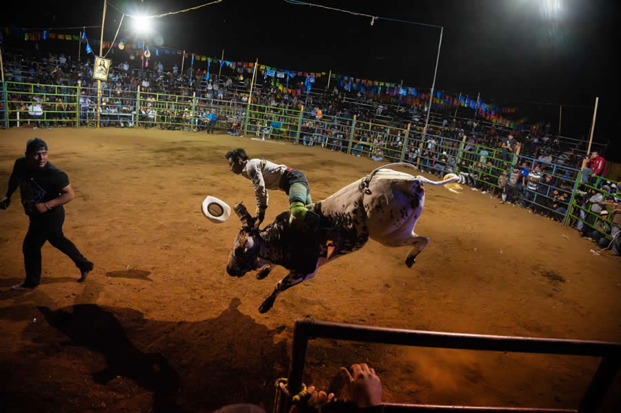 Bull rider thrown into the air during a rodeo at night, captured under bright lights with a crowd watching in the background, showing dynamic action and intense movement in a street-style documentary scene. - Mexico Street Photography by Tavepong Pratoomwong
