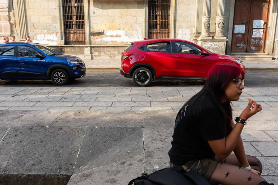 Woman with red hair sitting on a sidewalk eating, with red and blue cars parked behind her, creating a colorful and balanced street photography composition with urban textures. - Mexico Street Photography by Tavepong Pratoomwong