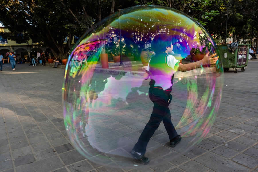 Child walking inside a large colorful soap bubble in a public park, with rainbow reflections surrounding him, creating a dreamy and playful street photography scene with vibrant colors and motion. - Mexico Street Photography by Tavepong Pratoomwong