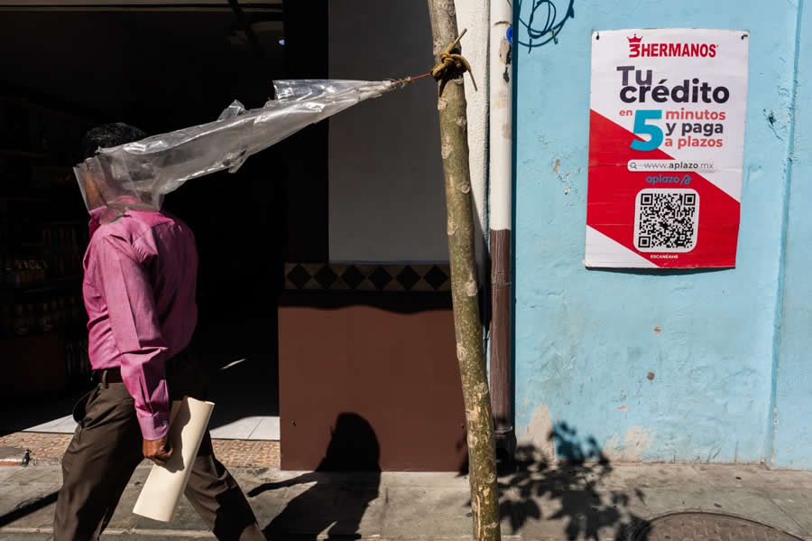 Man walking on a street with a plastic sheet stretched over his head, attached to a pole, creating a humorous and surreal street photography scene with strong shadows and contrasting colors. - Mexico Street Photography by Tavepong Pratoomwong