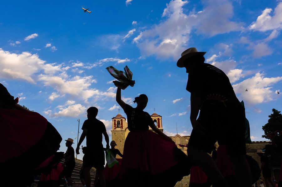 Silhouetted people dancing in traditional attire against a bright blue sky with clouds, with a historic building in the background, captured in a dynamic and layered street photography scene. - Mexico Street Photography by Tavepong Pratoomwong