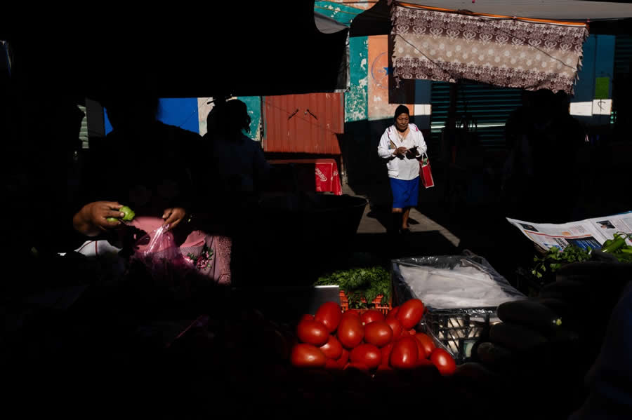 Market scene with strong shadows and a woman walking through a beam of light, surrounded by vegetables like tomatoes in the foreground, creating a high-contrast and layered street photography composition. - Mexico Street Photography by Tavepong Pratoomwong