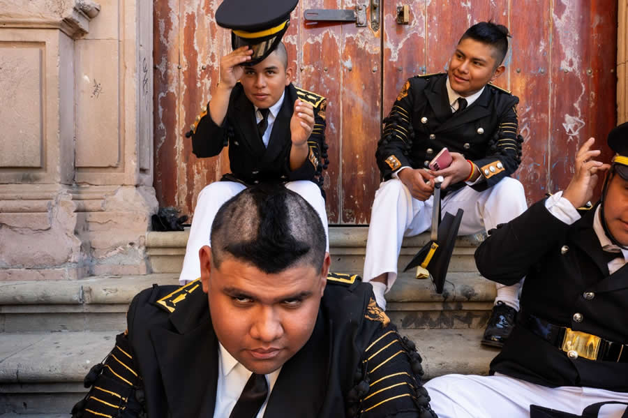 Group of uniformed men sitting on steps, adjusting hats and relaxing, captured in a candid street photography moment with textured wooden doors in the background and strong composition focusing on expressions and interaction. - Mexico Street Photography by Tavepong Pratoomwong