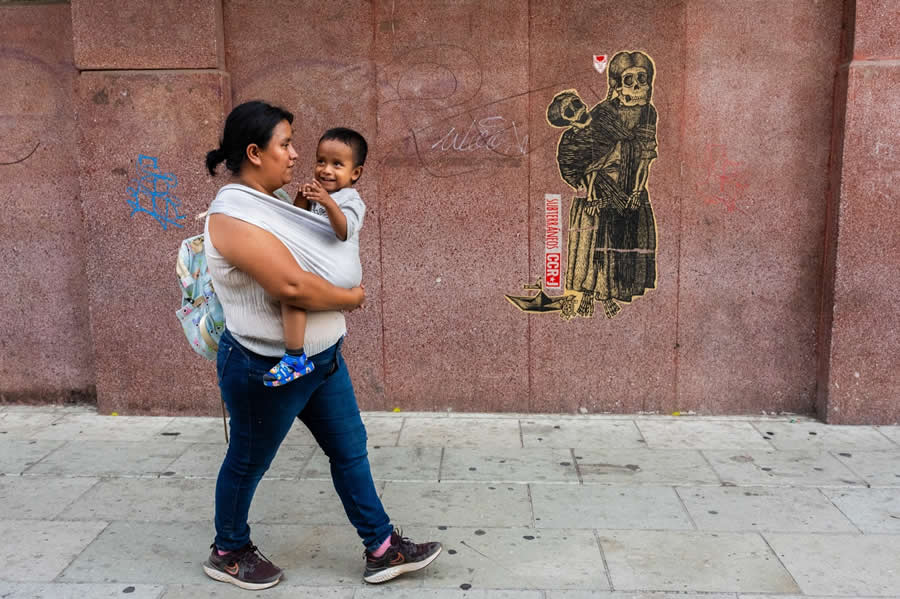 Woman carrying a child walking past a wall with a skeleton mural, creating a strong juxtaposition between life and death in a clean and well-composed street photography scene. - Mexico Street Photography by Tavepong Pratoomwong
