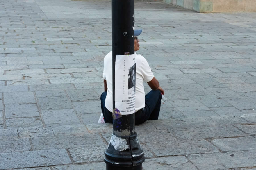 Man sitting on the ground partially hidden behind a lamppost, with only part of his face visible, captured in a minimalist and cleverly composed street photography scene with strong symmetry and visual humor. - Mexico Street Photography by Tavepong Pratoomwong