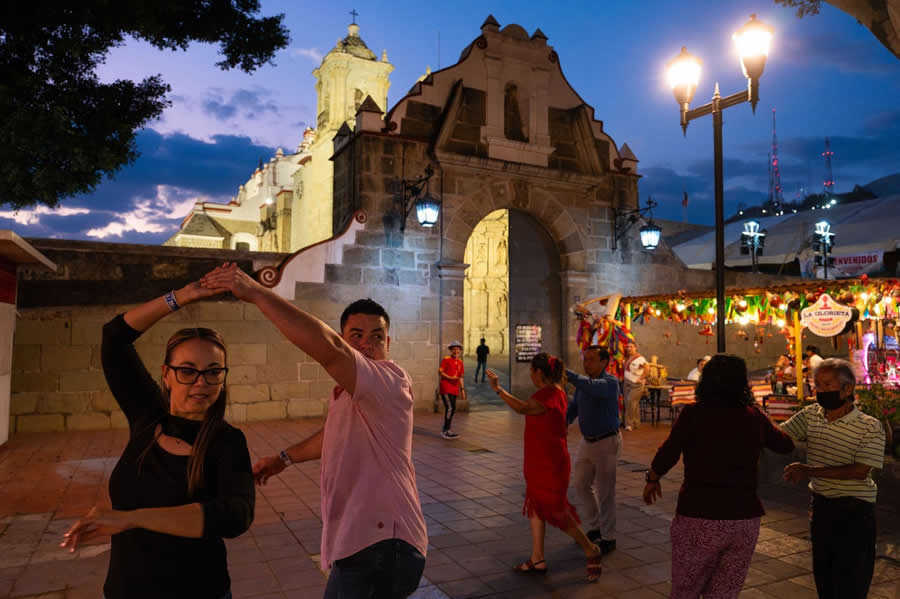 People dancing in a public square at dusk with historic buildings and warm streetlights, capturing a lively cultural scene with movement, colorful lights, and a vibrant evening atmosphere. - Mexico Street Photography by Tavepong Pratoomwong