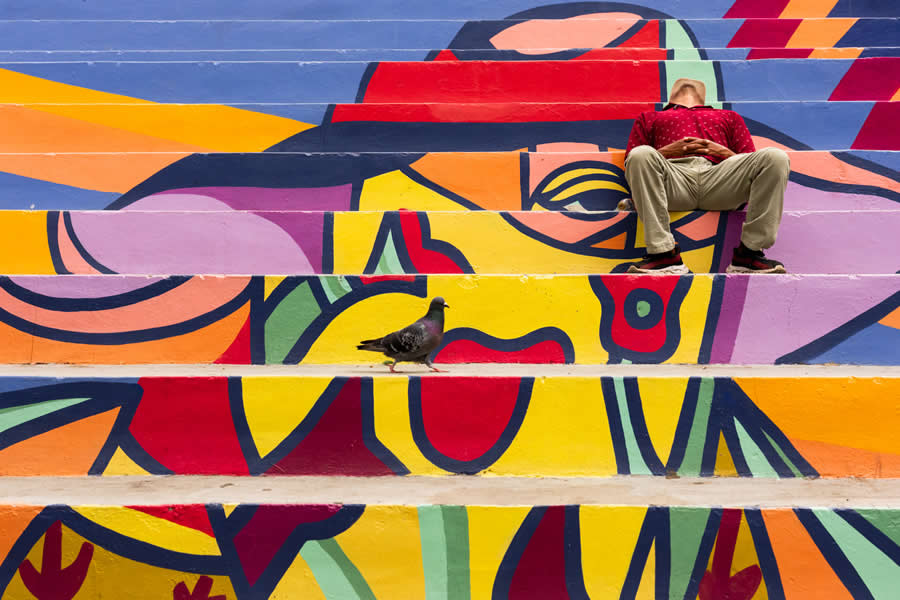 Man sitting on colorful painted steps with his head blending into a mural, creating a visual illusion, while a pigeon walks in the foreground in a vibrant and creative street photography composition. - Mexico Street Photography by Tavepong Pratoomwong