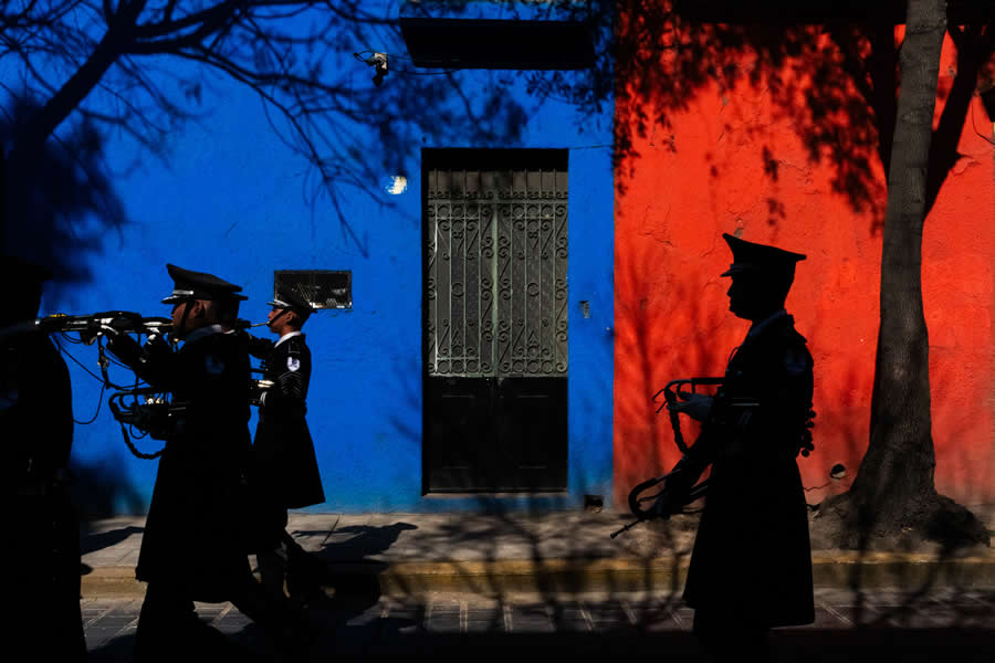 Silhouetted marching band members walking past a wall divided into blue and red colors, with strong shadows and contrast creating a bold and graphic street photography scene. - Mexico Street Photography by Tavepong Pratoomwong