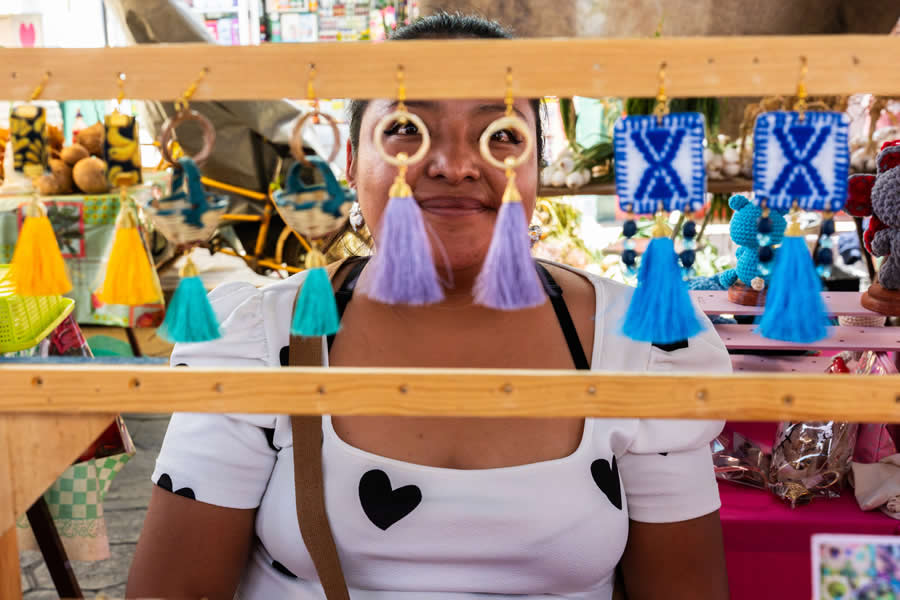 Smiling woman framed by colorful hanging tassels at a market stall, creating a vibrant and layered street photography portrait with strong use of foreground framing and bright colors. - Mexico Street Photography by Tavepong Pratoomwong