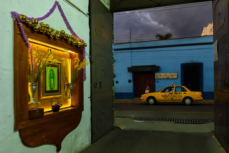 Indoor shrine with warm lighting on the left framed by an open doorway, revealing a yellow taxi passing on the street outside, creating a layered composition with contrast between interior and exterior scenes. - Mexico Street Photography by Tavepong Pratoomwong
