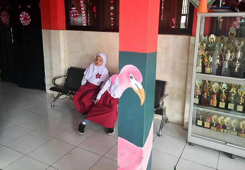 Street photo of a child sitting beside a pillar painted with a flamingo, creating an illusion where the bird aligns with the subject’s body.
