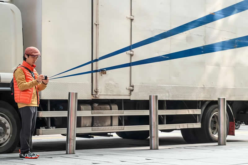 Street photo of a person holding a camera aligned with blue stripes on a truck, creating an illusion that the lines extend from the camera lens.