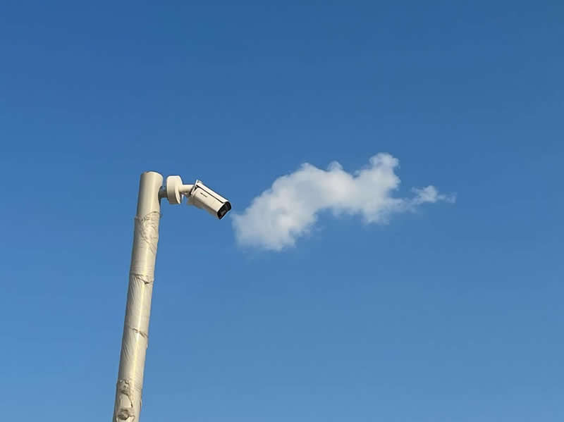 Street photo of a surveillance camera aligned with a cloud in the sky, creating the illusion that the camera is blowing out smoke.