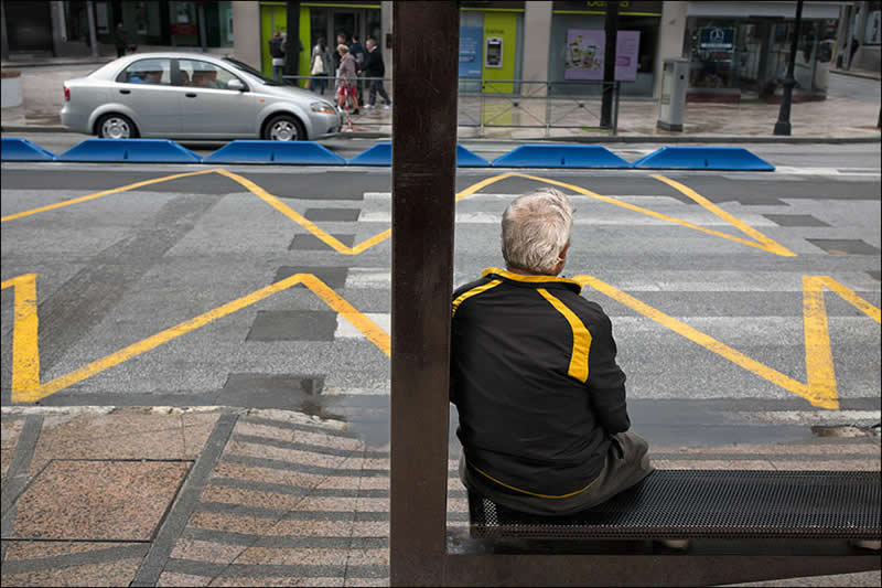 Street photo of a man sitting on a bench where yellow road lines align with the yellow stripes on his shirt, creating visual symmetry.