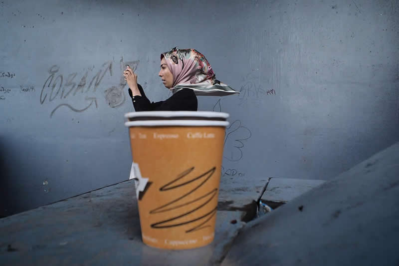 A woman writing on a wall appears aligned behind a large coffee cup in the foreground, creating a layered perspective illusion.