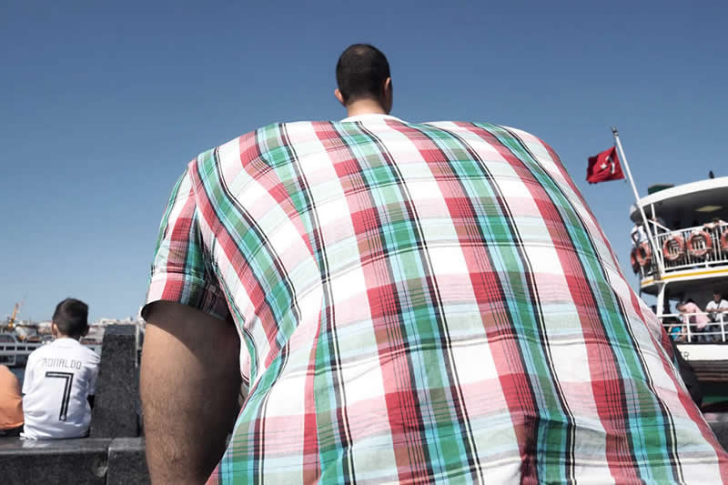 A man seen from behind appears extremely wide due to perspective, with a plaid shirt exaggerated by close framing in a street scene.