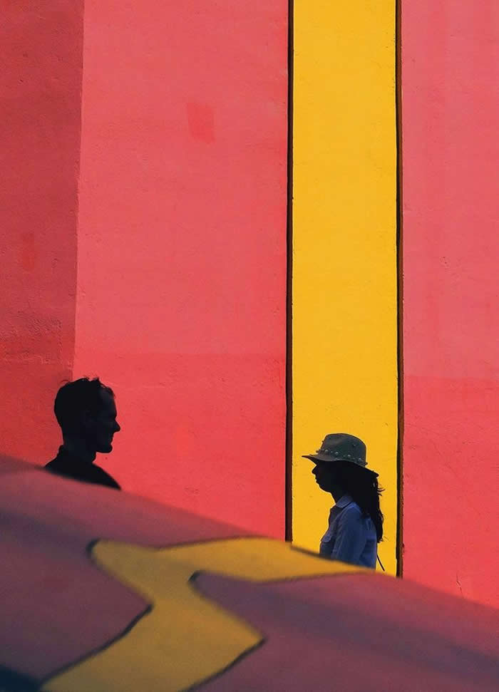 Silhouettes of two people facing each other against a vibrant red wall with a bold vertical yellow stripe, creating a minimalist geometric composition with strong contrast and color.