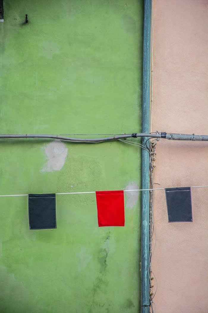 A minimalist scene of a green and beige wall divided by pipes, with three square cloths hanging on a line&mdash;two dark and one bright red&mdash;creating a striking contrast and geometric balance.