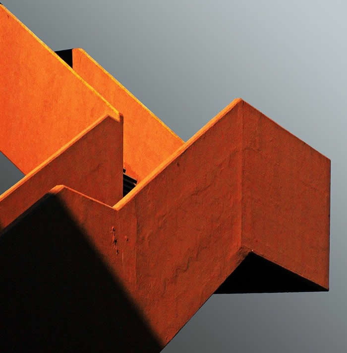 A minimalist architectural composition featuring sharp, angular orange building structures against a muted gray sky, with strong light and shadow emphasizing geometric forms and contrast.