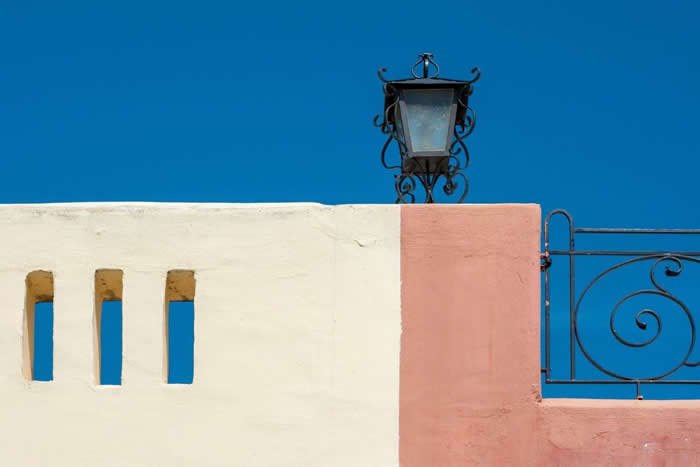 A minimalist architectural scene with pastel cream and pink walls, a decorative black lamp, and a clear blue sky, forming clean geometric shapes and balanced composition.