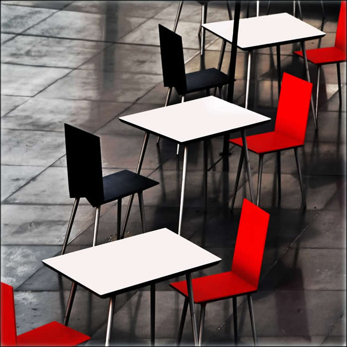 A minimalist arrangement of empty caf&eacute; tables with black and red chairs on a tiled floor, creating a rhythmic geometric pattern with strong contrast and repetition.