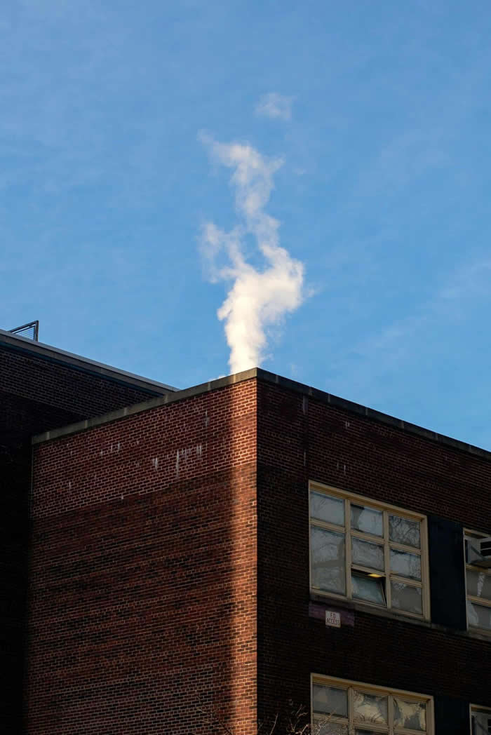 A minimalist urban scene showing a brick building corner under a clear blue sky, with white steam rising from the rooftop, creating a soft, cloud-like form against the geometric structure.