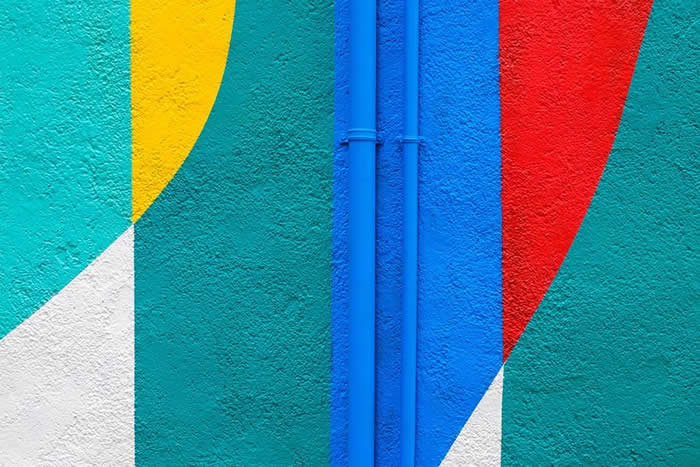 A vibrant abstract wall featuring curved geometric shapes in yellow, red, white, and teal, intersected by two vertical blue pipes, creating a bold minimalist composition of color and symmetry.