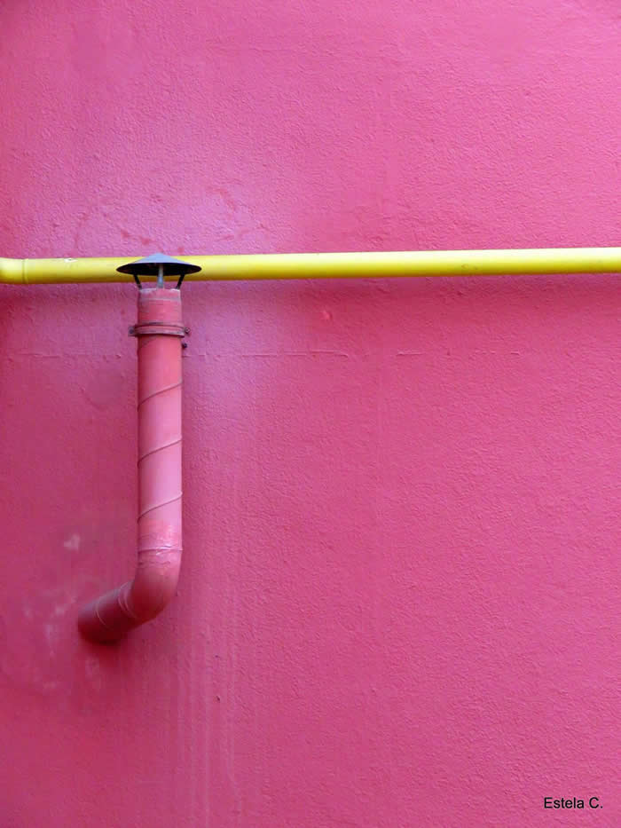 A minimalist composition of a pink wall intersected by a bright yellow horizontal pipe and a small vertical red pipe, creating a simple yet striking geometric contrast.