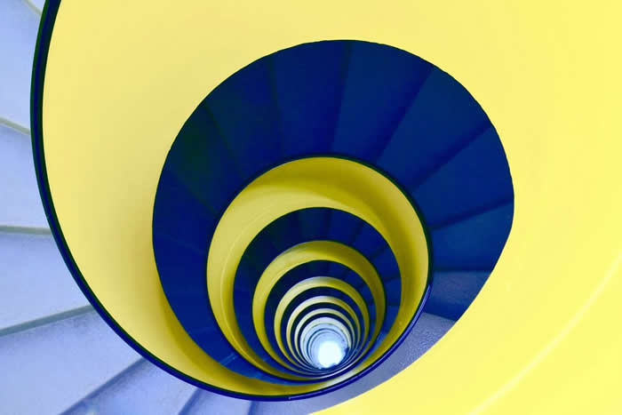 A minimalist view of a spiral staircase in bold yellow and deep blue tones, forming concentric circular patterns that lead the eye toward a bright central vanishing point.