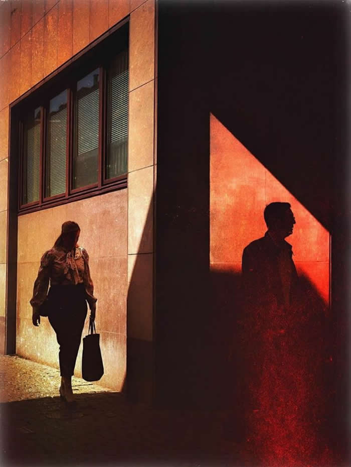 A dramatic street scene showing a woman walking in sunlight beside a building, while a man&rsquo;s silhouette appears in a sharply lit red triangular shadow, creating contrast between light, shadow, and human presence.