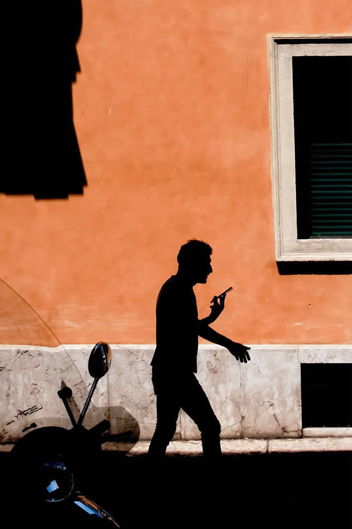 A silhouette of a man walking while looking at his phone against a warm orange wall, with a window and strong shadows creating a minimalist urban composition.