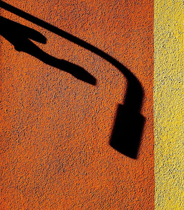 A minimalist composition of an orange textured surface with a yellow edge, featuring the shadow of a hand reaching toward a curved streetlight shadow, creating a dramatic interplay of shapes and contrast.
