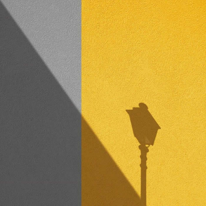 A minimalist composition of a wall split between gray and yellow, with a sharp diagonal shadow and the silhouette of a streetlamp creating clean geometric contrast.