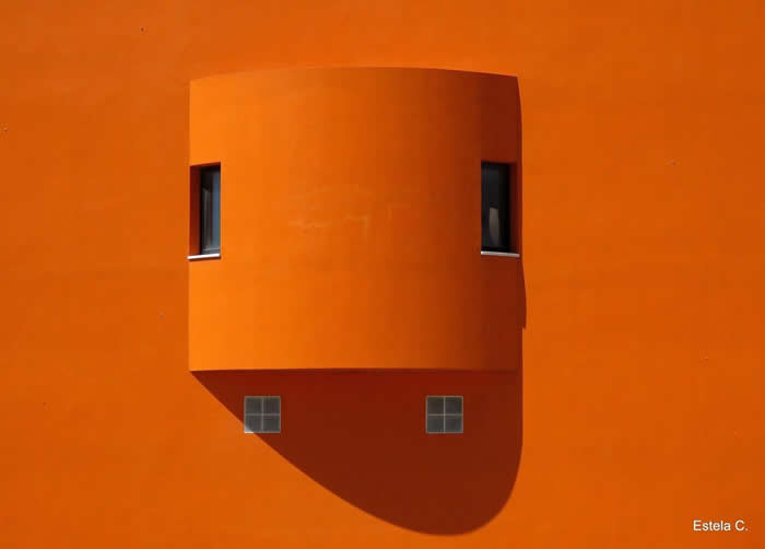 A minimalist orange facade featuring a protruding cylindrical structure with two small windows, casting a soft curved shadow that emphasizes clean geometry and bold monochromatic color.