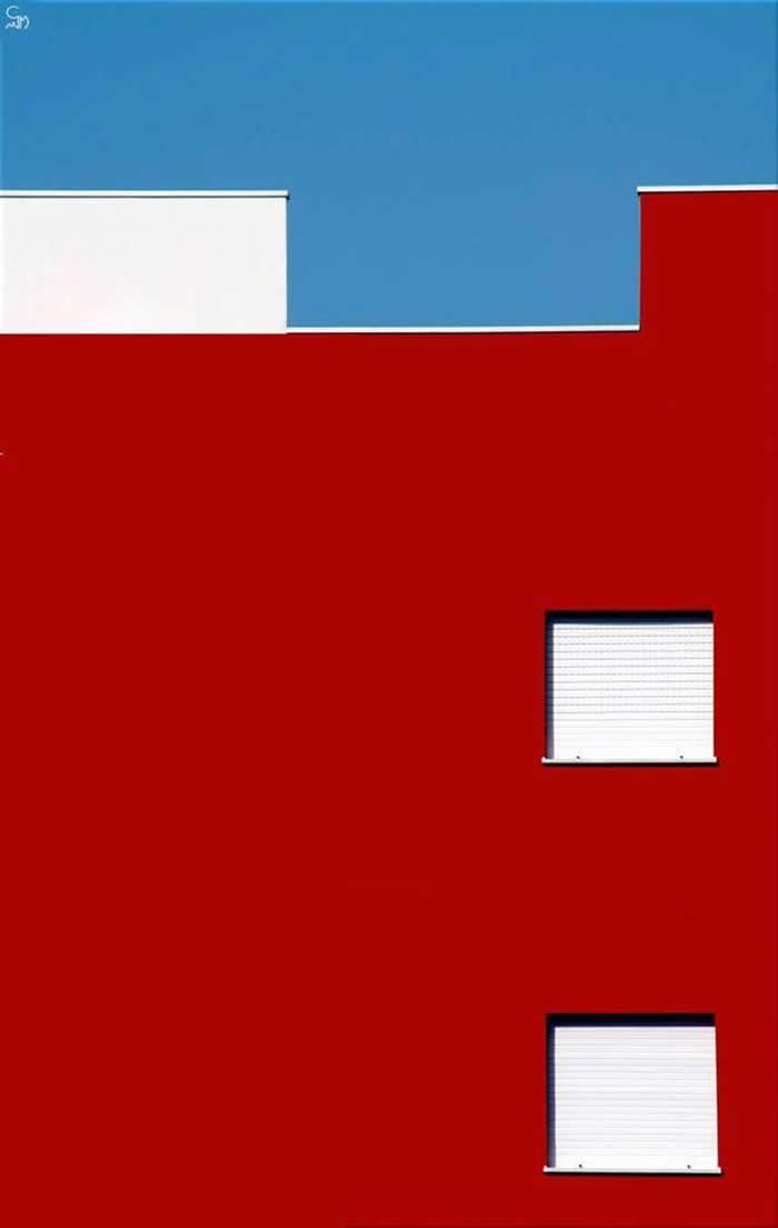 A minimalist architectural composition featuring a bold red building facade with two small white windows and a stepped white roofline against a clear blue sky, forming clean geometric shapes and strong color contrast.