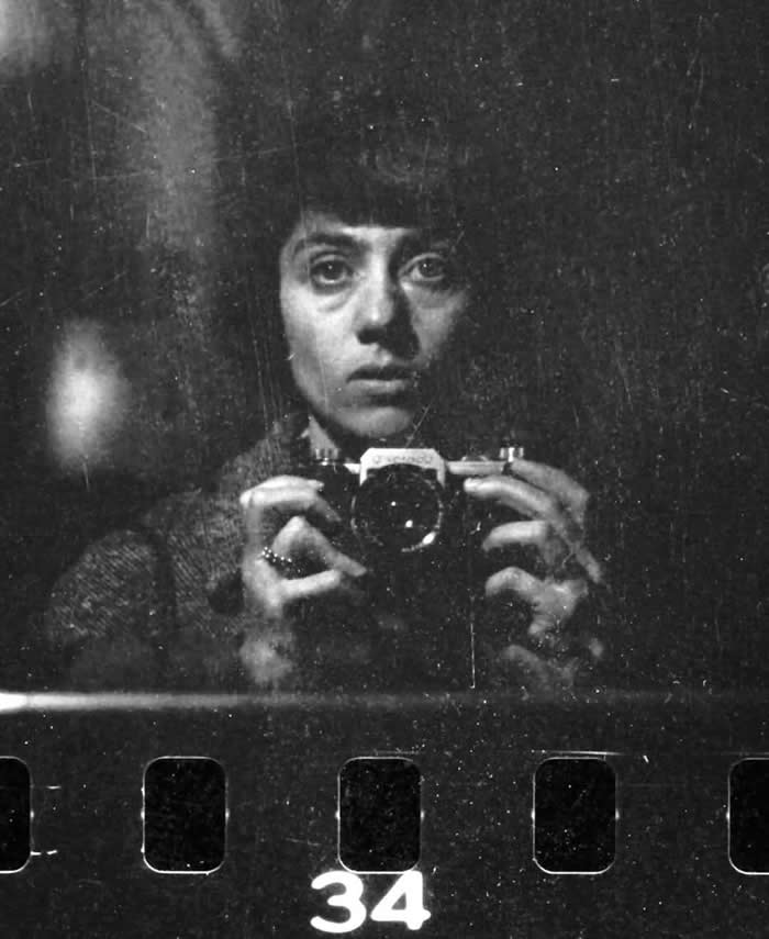 Black-and-white mirror self-portrait of photographer Diane Arbus holding a camera close to her face, staring directly into the lens, with visible film negative frame markings at the bottom.