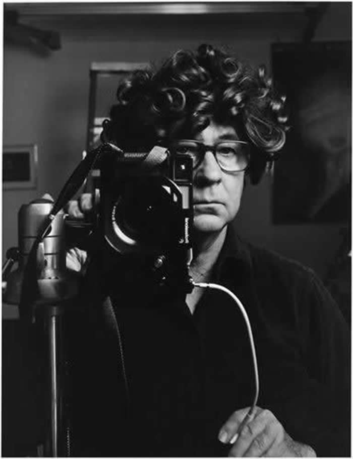 Black-and-white mirror self-portrait of photographer Elliott Erwitt wearing hair curlers and glasses, standing beside a camera on a tripod, looking directly into the mirror.