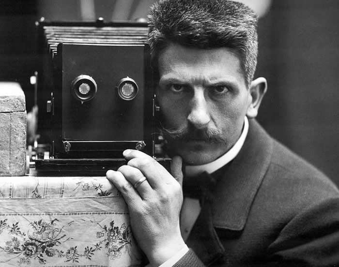 Black-and-white self-portrait of photographer Fr&eacute;d&eacute;ric Boissonnas posing beside a vintage box camera, staring intensely toward the viewer, with one hand resting near the camera.