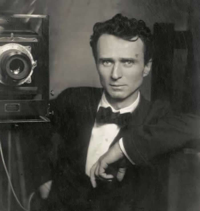 Black-and-white self-portrait of photographer Edward Steichen in formal attire with a bow tie, posing beside a large vintage camera and looking directly at the viewer.