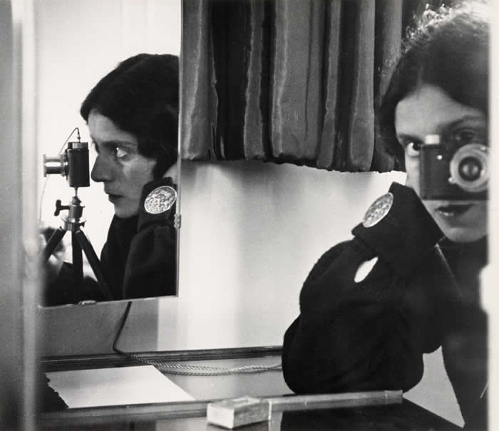 lack-and-white mirror self-portrait of photographer Ilse Bing holding a camera close to her face, with her side profile reflected in a second mirror beside her.
