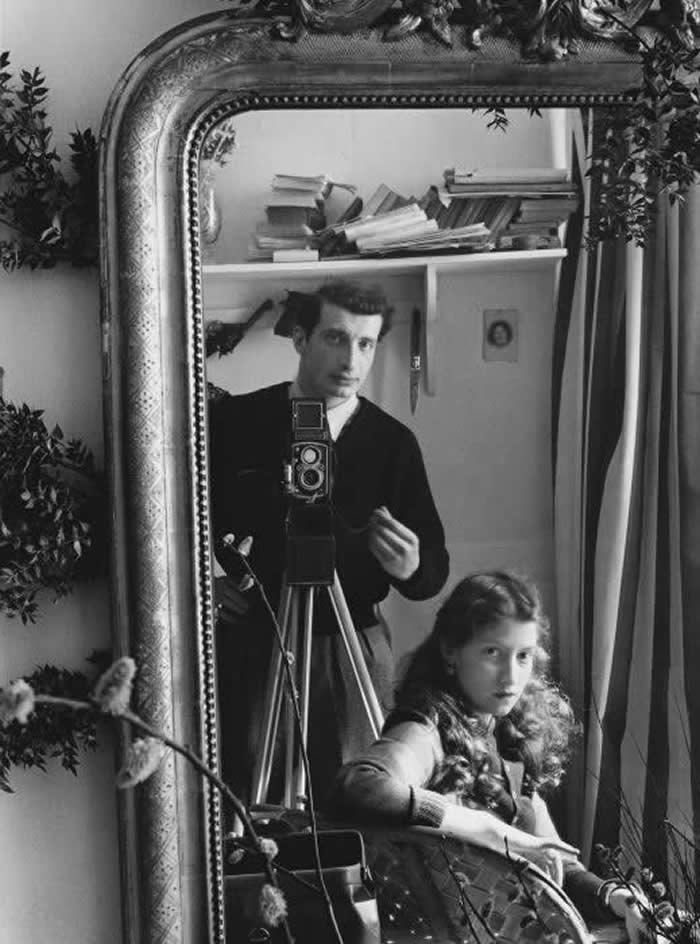 Black-and-white mirror self-portrait of photographer &Eacute;douard Boubat standing behind a camera on a tripod, with companion Lella seated in front, reflected in an ornate framed mirror.
